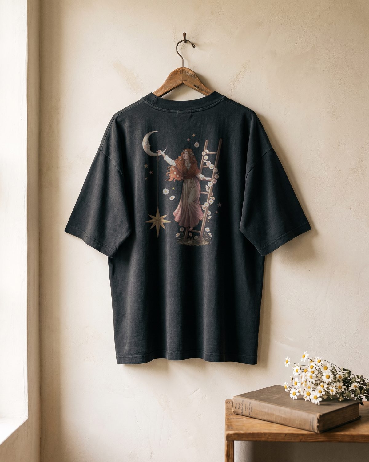 Mockup of the back of a faded black oversized tee hanging on a wall. The print shows the copper-haired woman standing on a ladder of daisies, hanging a gold star from the horn of the crescent moon.