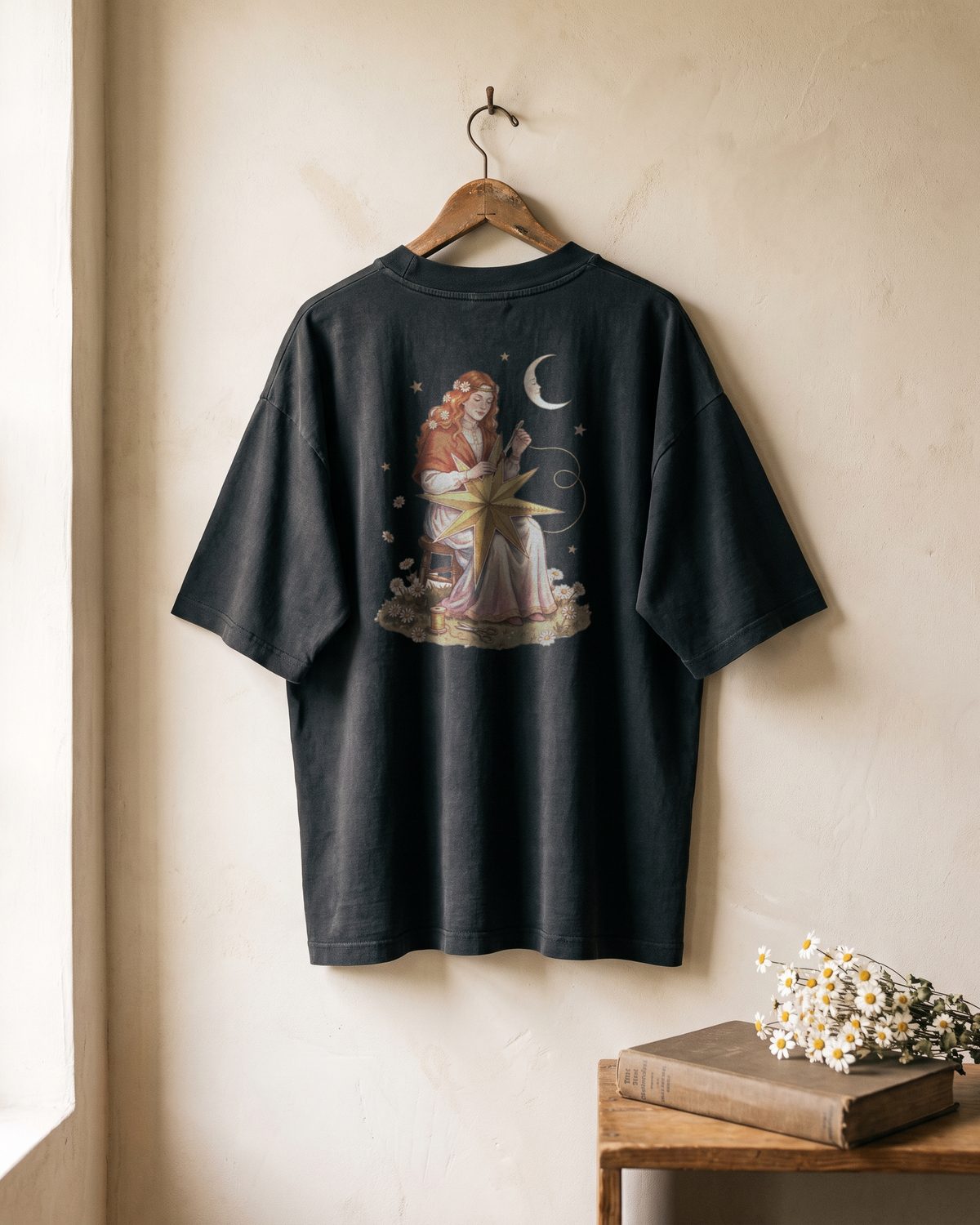 Mockup of the back of a faded black oversized tee hanging on a wall. The print shows the copper-haired woman sewing a torn gold star with gold thread, a crescent moon beside her.